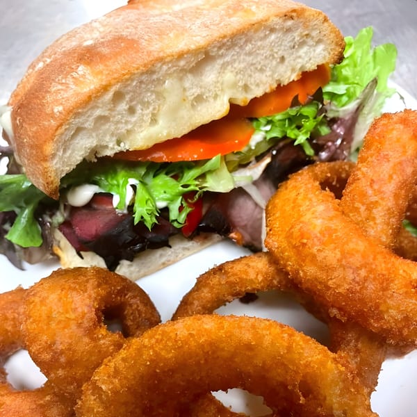 A roast beef sandwich on ciabatta with lettuce, tomato, and mayo, served with onion rings.